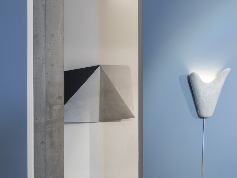 Geometric wall sculpture beside a textured, white wall-mounted light fixture against a blue wall. Concrete pillar visible on the left.