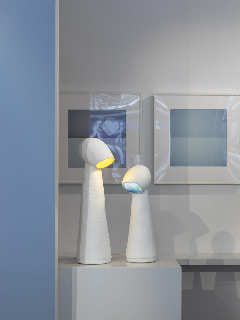 Two tall, white, modern lamps with curved shades on a pedestal. One emits a yellow light and the other a cool white light. Three framed artworks on a gray wall in the background.