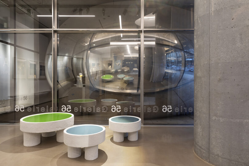 Modern gallery with reflective mirrored wall, showcasing abstract art. Three round, multicolored stools are in the foreground. Glass wall and concrete elements are visible.
