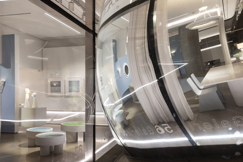 Modern art gallery with reflective, curved glass exterior. Interior features sculptures, framed artwork, and minimalist furniture in a softly lit setting.