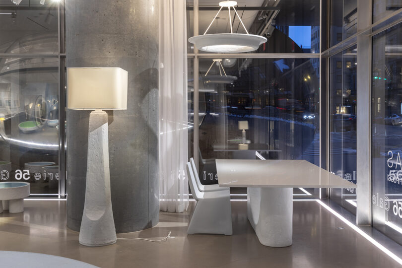 Modern interior with a white table and two chairs near large windows. A tall, sculptural lamp and a circular ceiling light illuminate the space. Concrete column and sheer curtains are visible.