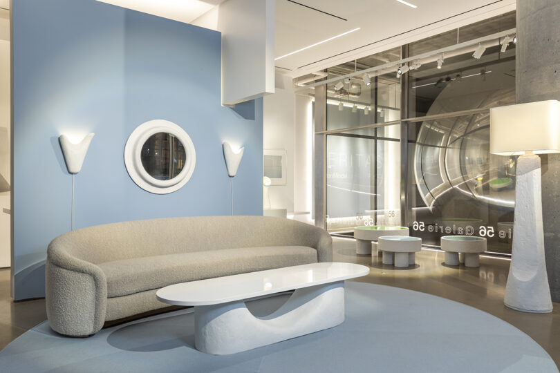 A modern showroom with a beige curved sofa, white oval coffee table, and unique wall and floor lamps. Blue and white color scheme with large glass windows and circular wall decoration.