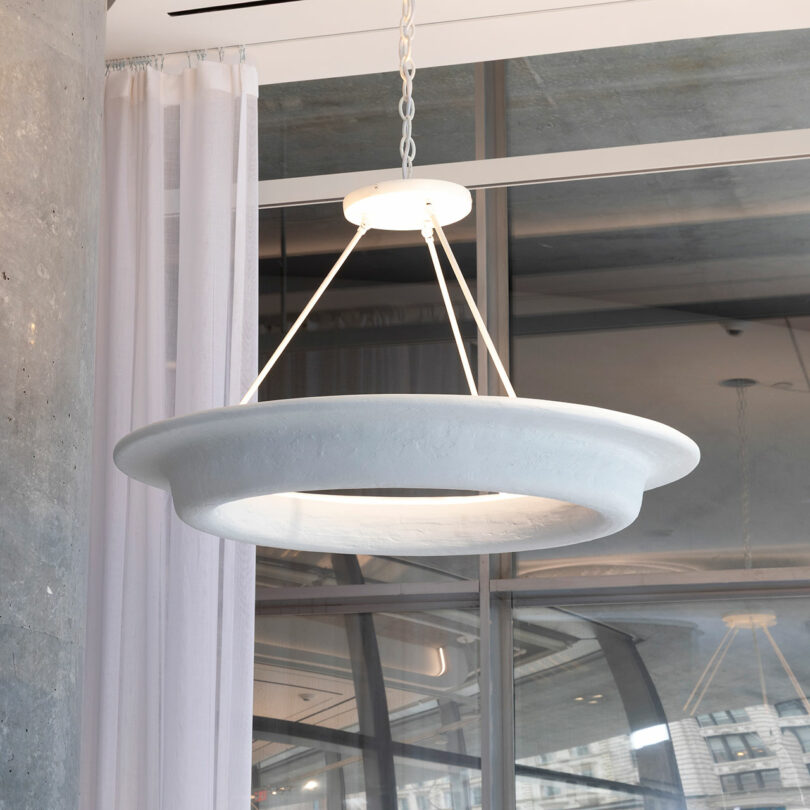 A modern, circular pendant light designed by Veritas hangs from the ceiling with a matte finish. Supported by four cables, it elegantly complements the backdrop of large windows and sheer curtains, echoing the sophisticated style of Lee F. Mindel.