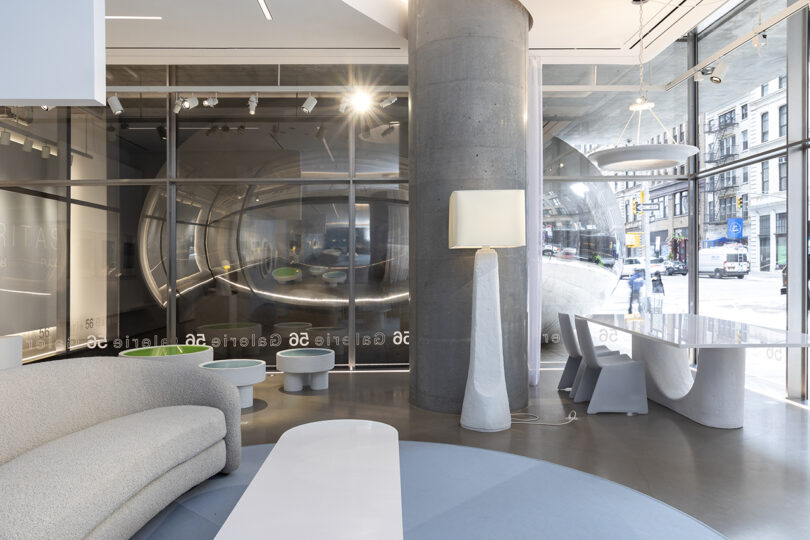 Modern showroom with sleek, curved furniture, large cylindrical sculpture visible through glass walls, and minimalistic decor with neutral tones.