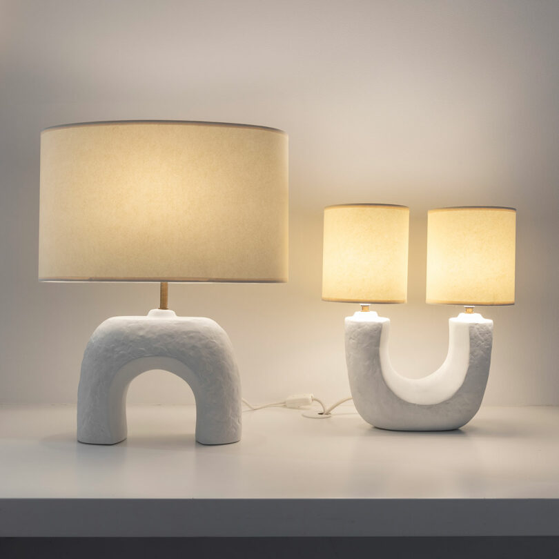 Two modern Veritas white ceramic table lamps with connected bases and beige cylindrical shades sit elegantly on a white surface, illuminated against a plain wall.