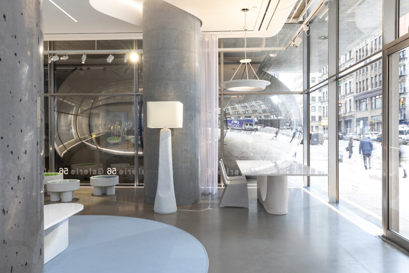 A modern office lobby with curved walls, a floor lamp, white tables, and large windows showing a city street reflection outside.