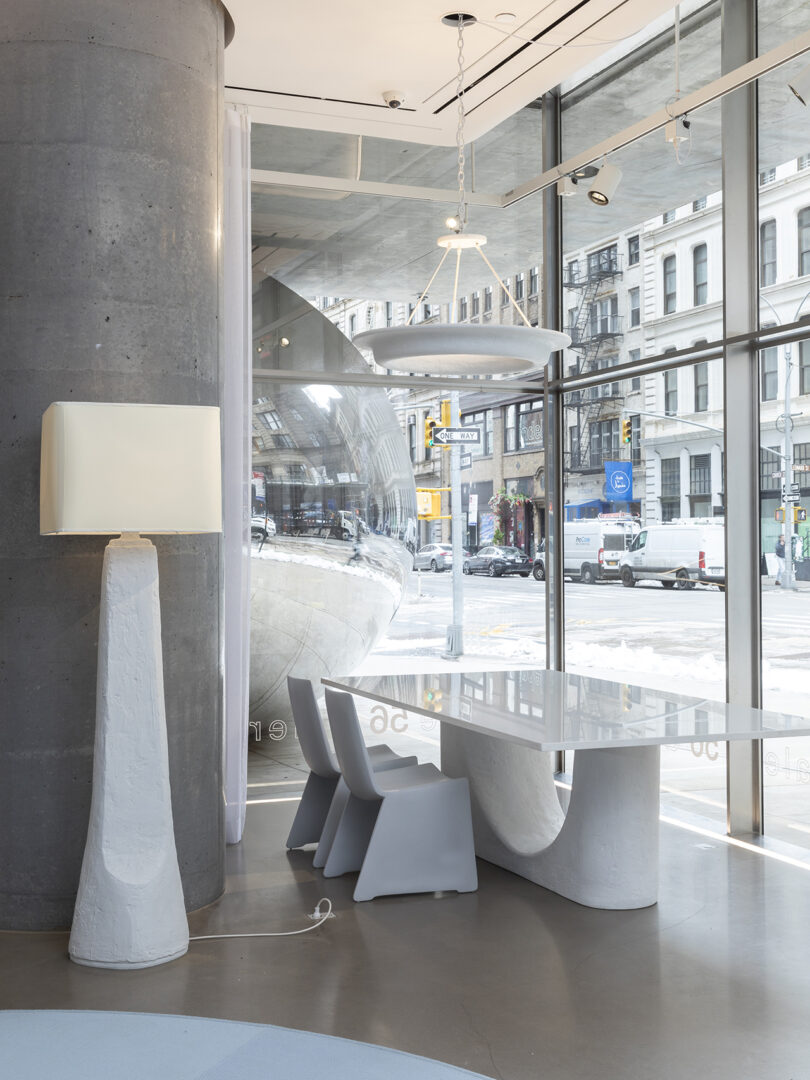 Modern interior with a sleek white table and chairs near large windows, featuring a unique tall lamp and urban street view outside.