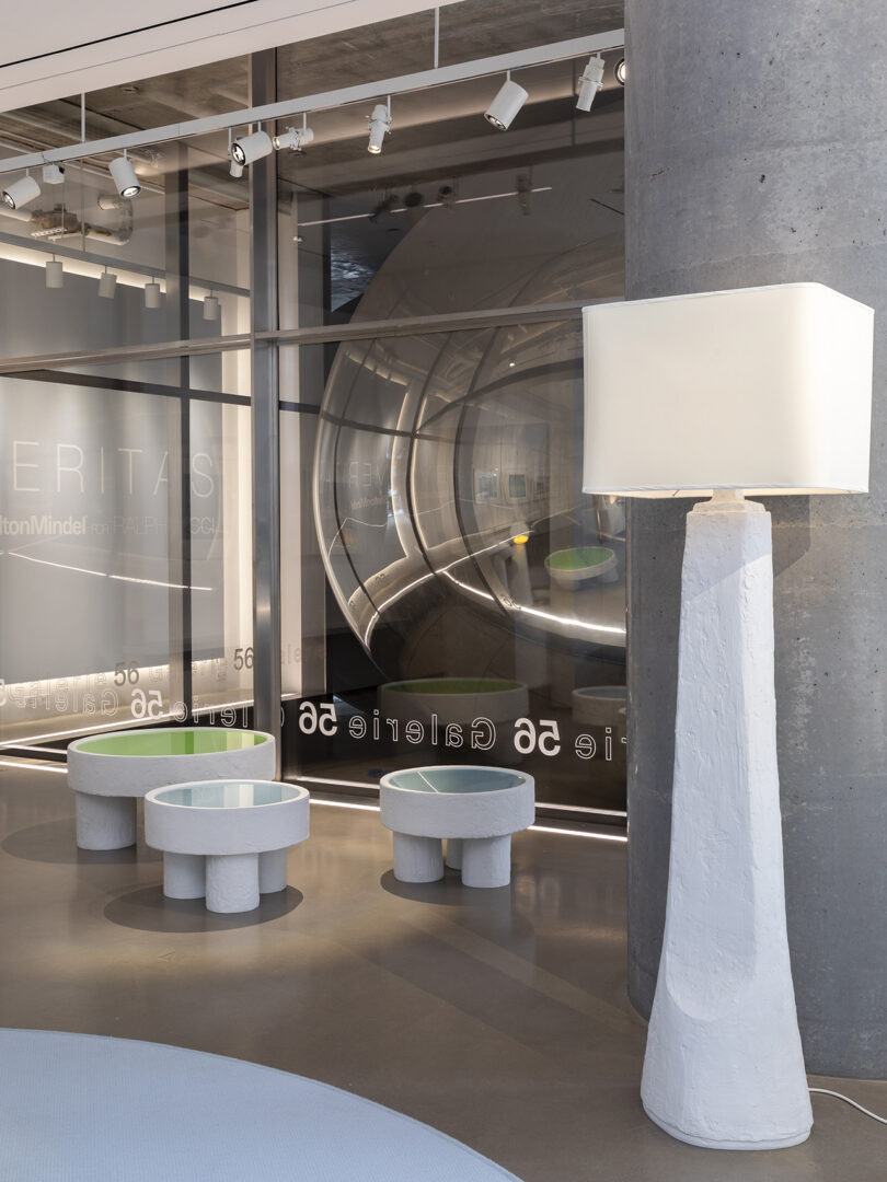 Modern interior with a tall, white floor lamp and three round, white tables with glass tops in front of a storefront window displaying the text 