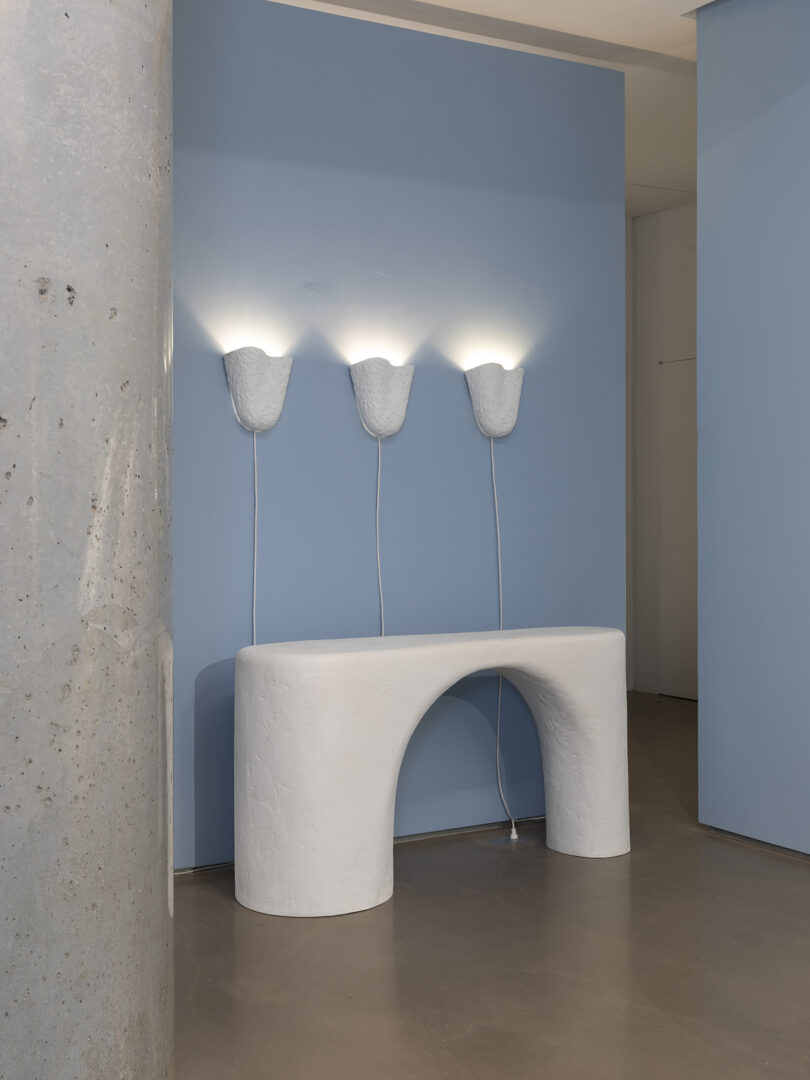 A minimalistic white console table with an arch stands against a blue wall, beneath three sculptural wall sconces casting light upwards. A concrete pillar is on the left.