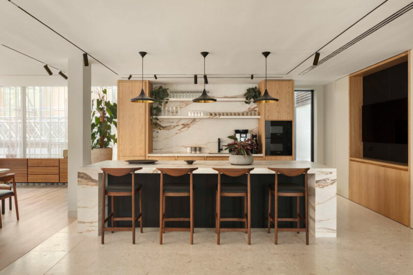 Modern kitchen with a marble island, four wooden barstools, pendant lights, and wooden cabinets. Plants and decor items are placed on shelves. Dining room visible in the background.