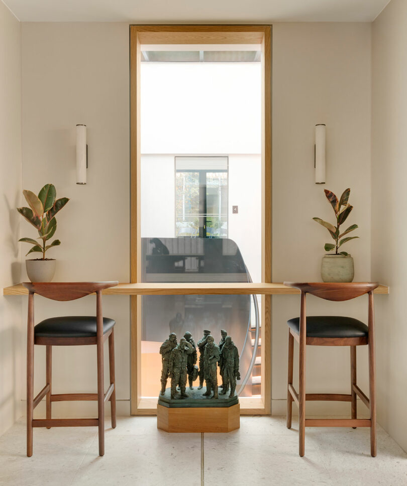 Two wooden chairs with black cushions at a narrow table, flanked by potted plants. Behind is a window displaying a modern interior staircase. A small sculpture sits below the table.