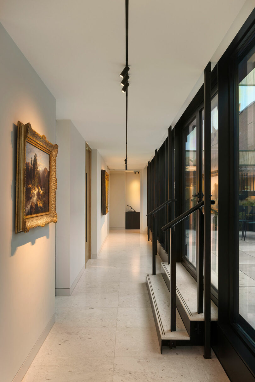 A modern corridor with beige floors, a row of large windows on the right, paintings on the left wall, and a small set of stairs leading down.