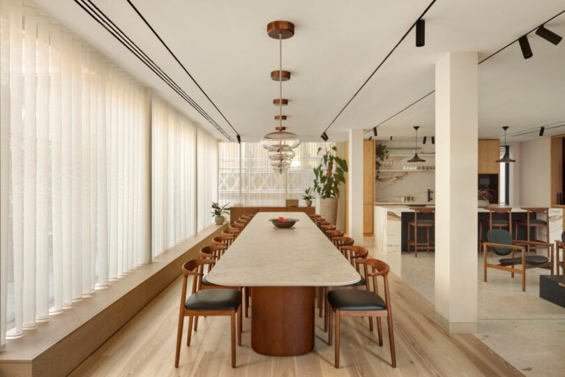 Modern dining room with a long table, wooden chairs, and pendant lights. Large windows with vertical blinds line one wall. The kitchen is visible in the background.