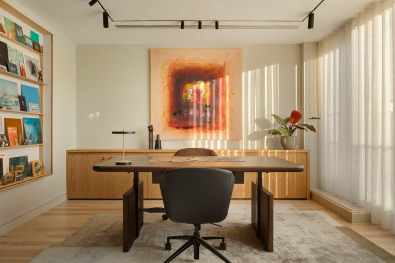Modern office with a wooden desk, black chair, large abstract painting, bookshelves, and a potted plant by floor-to-ceiling windows with sheer curtains.