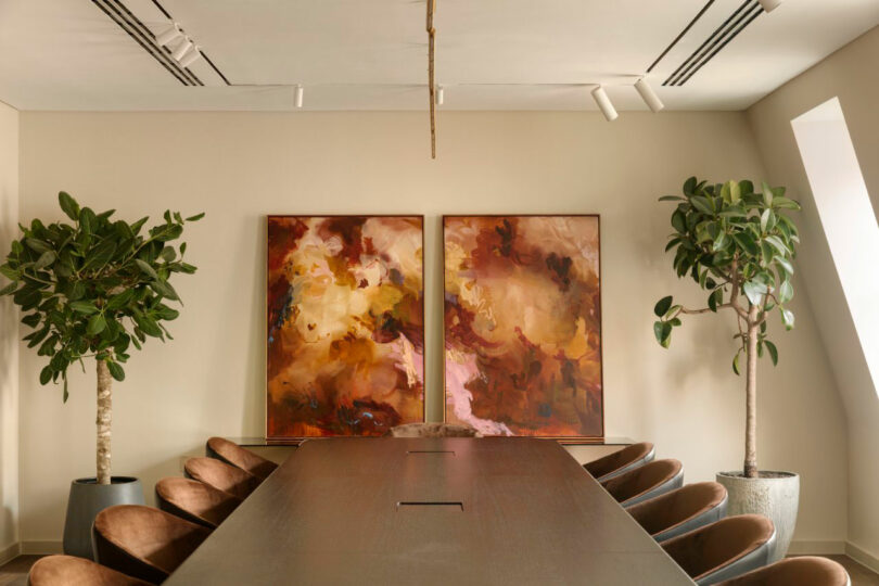 A conference room with a long table, brown chairs, abstract paintings on the wall, and potted plants on either side.