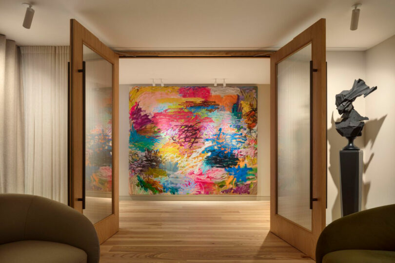 Open double doors reveal a colorful abstract painting on the wall, flanked by modern sculptures, in a well-lit room with beige chairs and wooden floors.