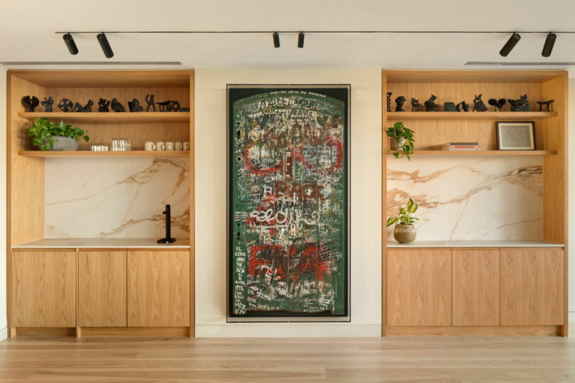 Wooden shelves with marble backs flank a central abstract painting. Decor includes plants and small sculptures. Track lighting illuminates the scene above.