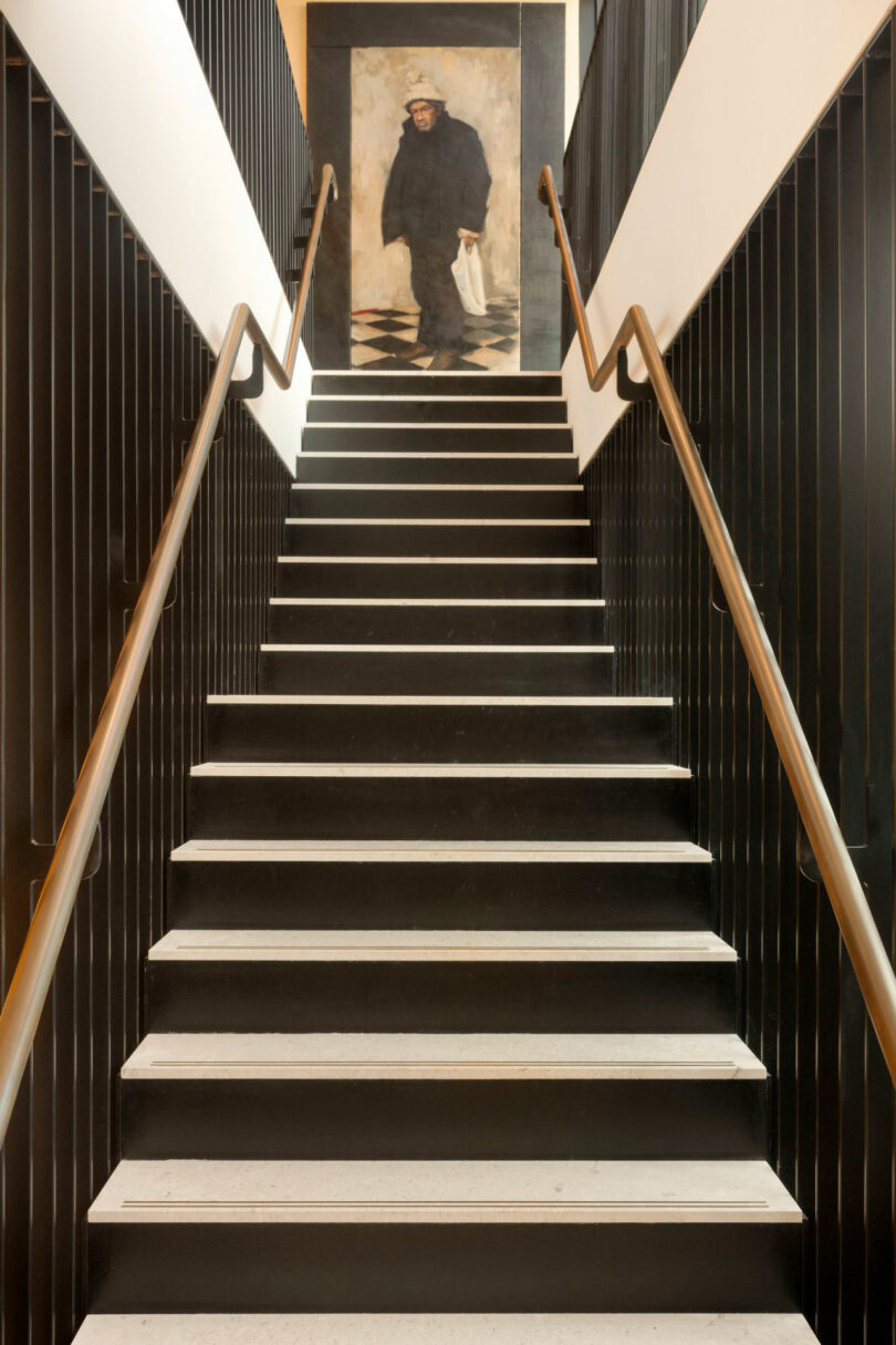 A staircase with black railings leads up to a landing, where a large painting of a person in dark clothing and a hat is displayed on the wall.