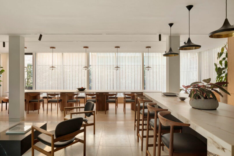 A modern dining area with wooden tables and chairs, pendant lights, and large windows with sheer curtains. A marble countertop with potted plants is at the front.