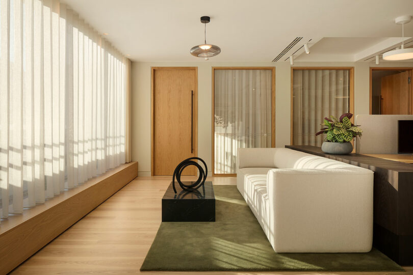 Modern office waiting area with a white couch, black sculpture on a dark pedestal, potted plant, and long curtains. Natural light highlights the wooden floor and accents.