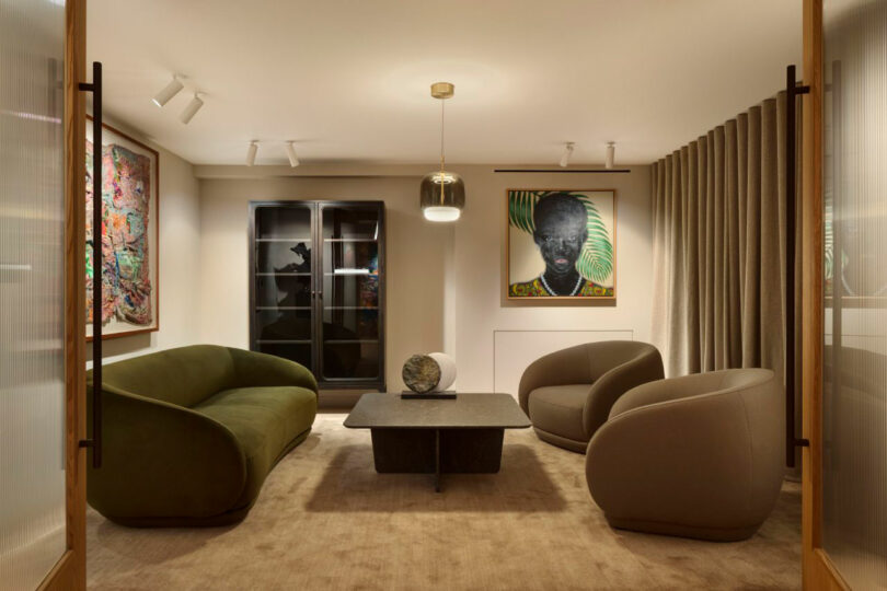 Modern living room with two armchairs, a sofa, and a central table. Artworks hang on the walls, and a glass cabinet is at the back. Ceiling lights illuminate the space.