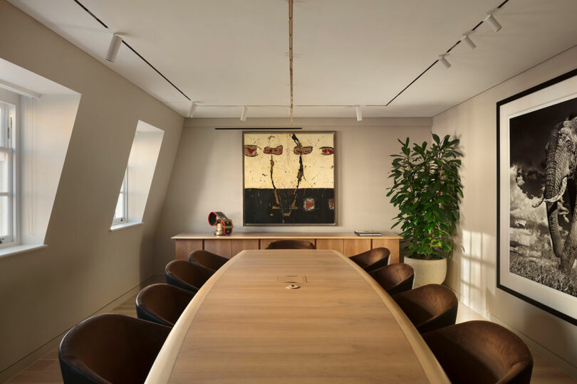 A conference room with a long wooden table, black chairs, an abstract painting, a large plant, and natural light from windows on the left.
