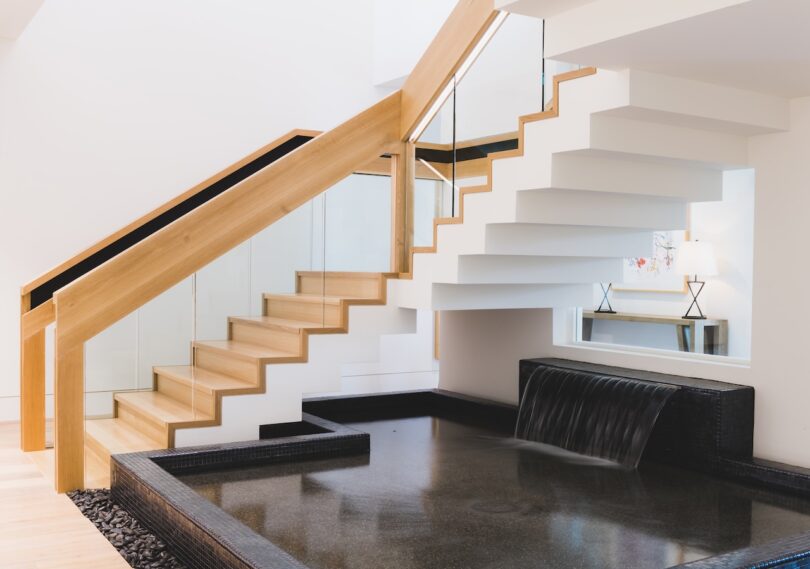 Modern interior with a wooden staircase, glass railing, and black stone water feature with a cascading waterfall. White walls and a console table with a lamp in the background.