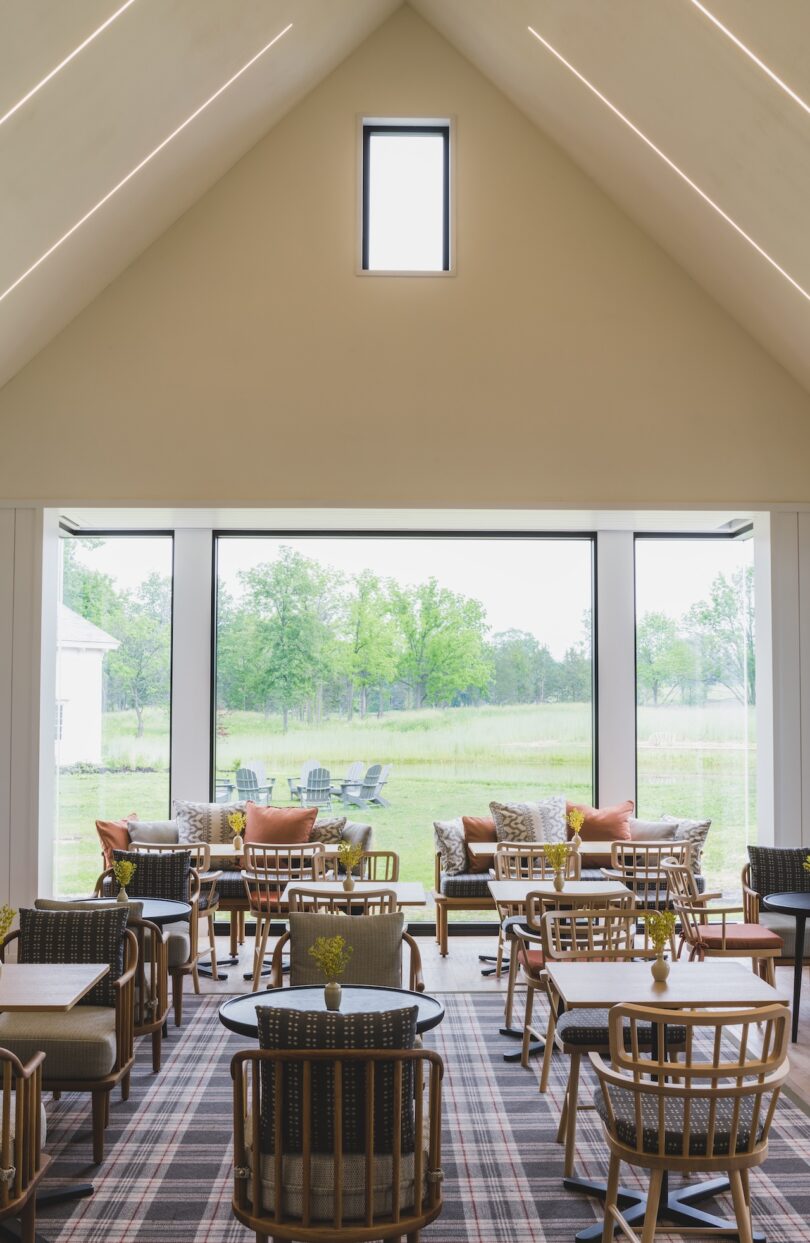Modern room with large windows, wooden chairs, and tables. Outside, greenery and trees are visible through the glass.