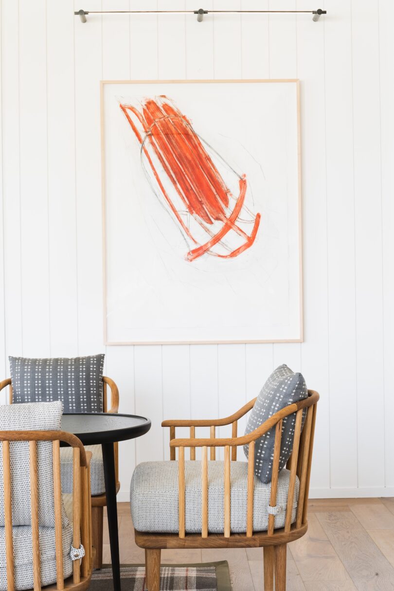 Two wooden chairs with gray cushions surround a round black table. Above, an abstract artwork with bold red strokes is displayed on a white paneled wall.