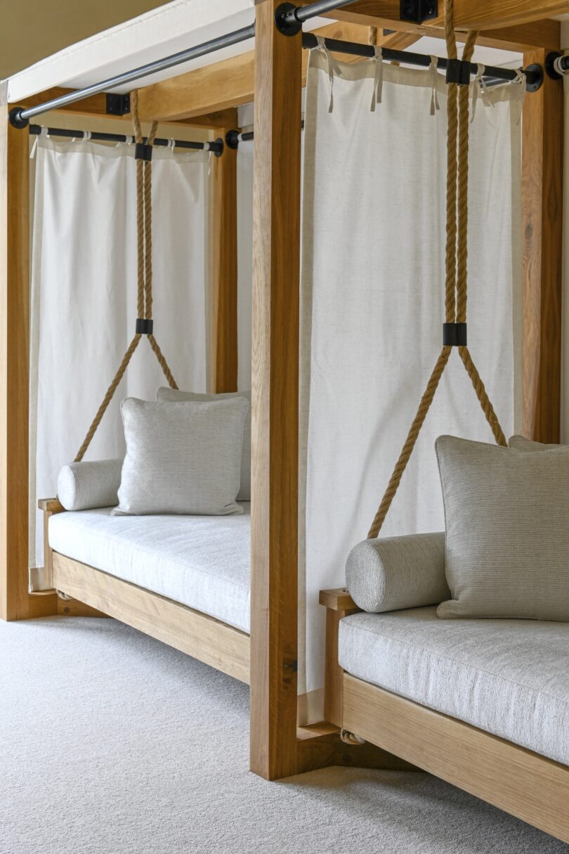 Two wooden hanging daybeds with cushions and white curtains, suspended by ropes, in a neutral-toned room.