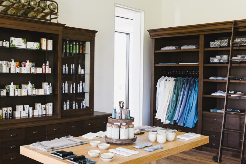 A store interior with wooden shelves displaying skincare products, neatly folded clothes, and ceramic cups on a central wooden table surrounded by neatly arranged items. A ladder leans on a shelf.