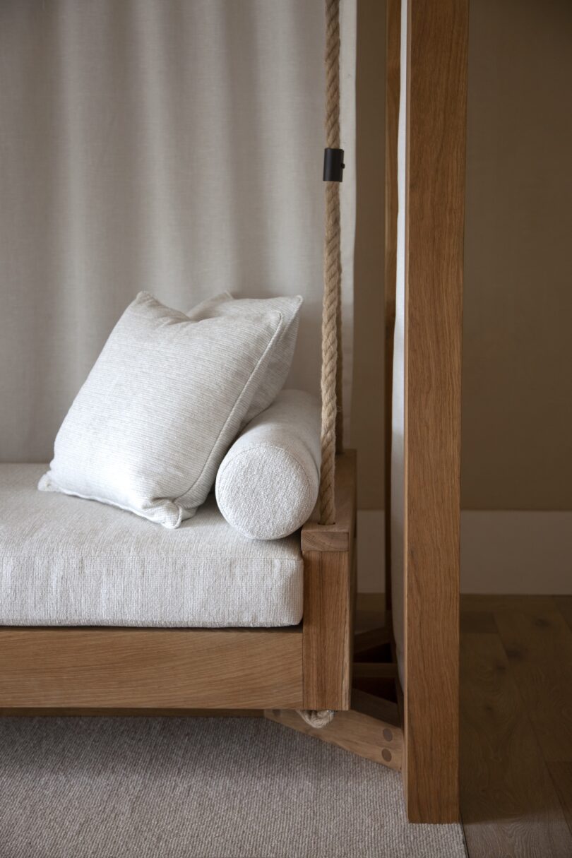 Wooden-framed swing with light gray cushions, including a square and cylindrical pillow, hangs indoors on thick ropes over a textured rug.
