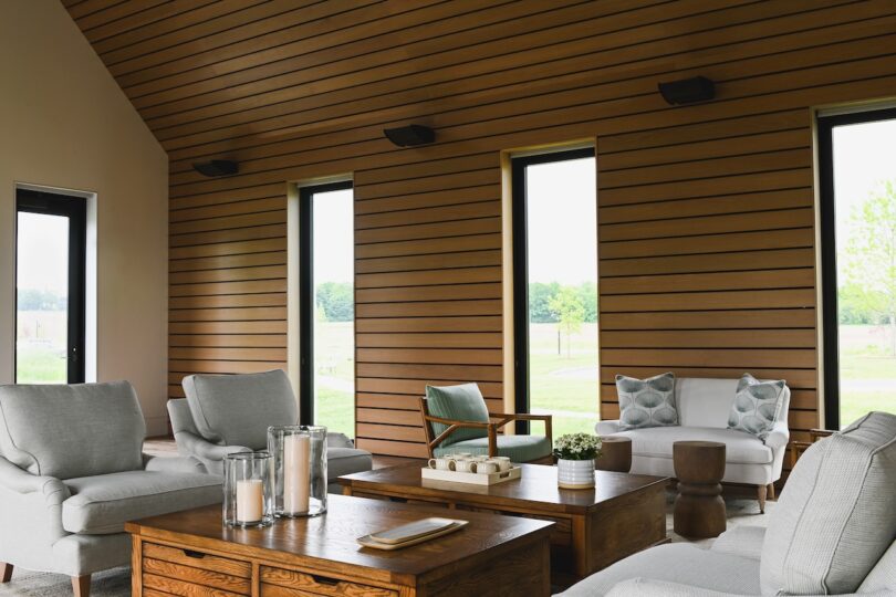Spacious modern living room with wood-paneled walls, large windows, gray armchairs, and a wooden coffee table adorned with candles.