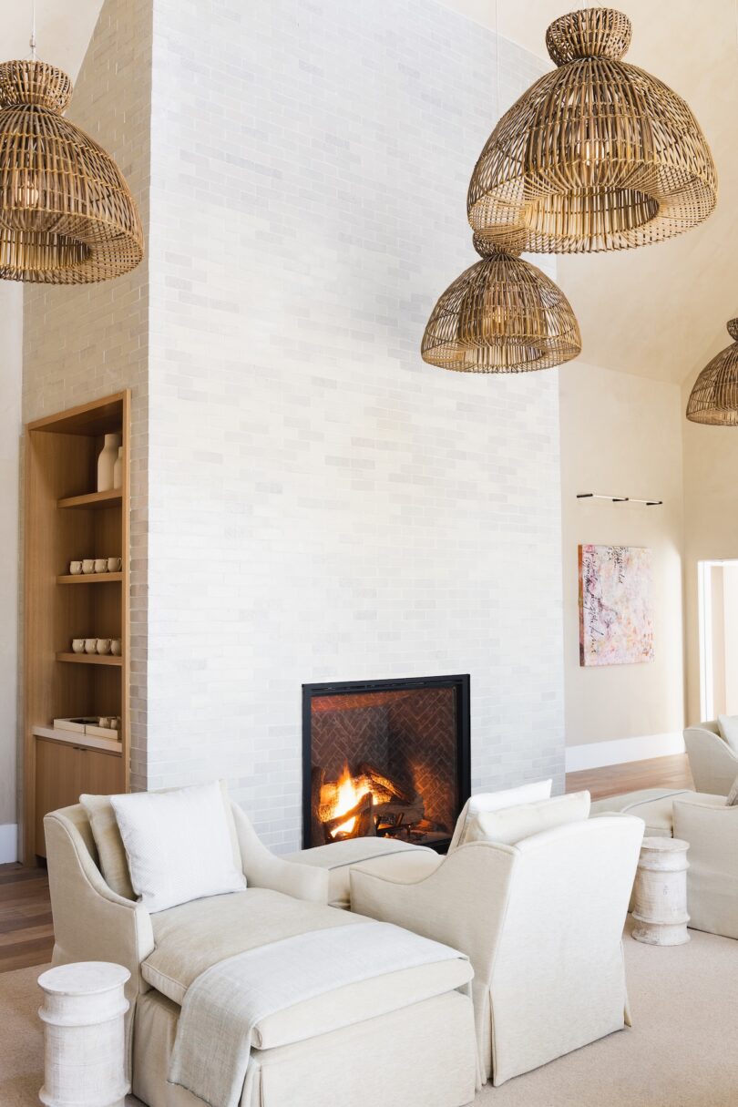 A modern living room with a lit fireplace, wicker pendant lights, beige armchairs with footrests, and shelves with vases. A textured wall and abstract art piece are also visible.