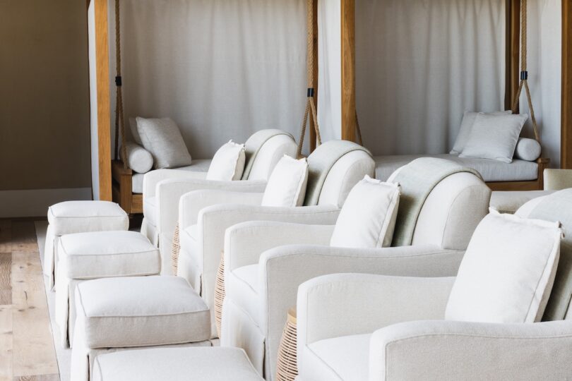 A row of white cushioned lounge chairs with matching ottomans is lined up in a well-lit room with wooden flooring and curtained daybeds in the background.