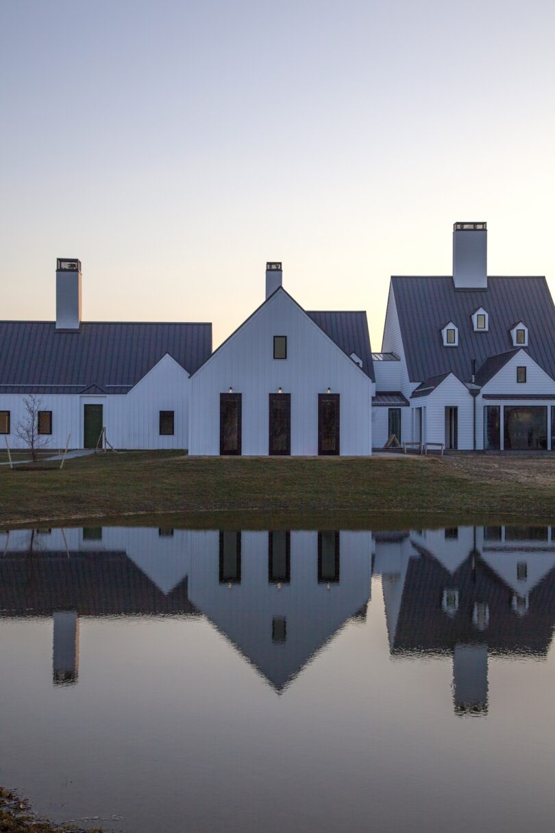 A row of white houses with steep roofs is reflected in a calm body of water, under a pale evening sky.