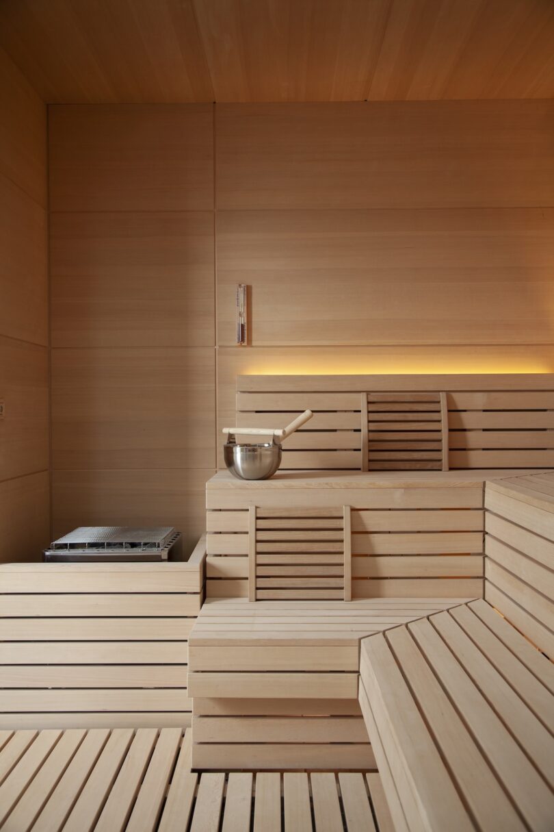 A wooden sauna interior with tiered seating, a metal bucket, a ladle, and a heater on the left. Warm lighting enhances the smooth wood panels.