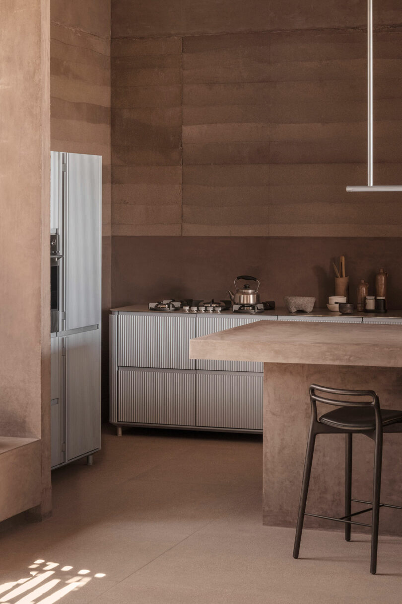 Modern kitchen with brown-toned walls, a metal fridge, stove, and a countertop with stools. Minimalist decor and soft lighting create a warm ambiance.