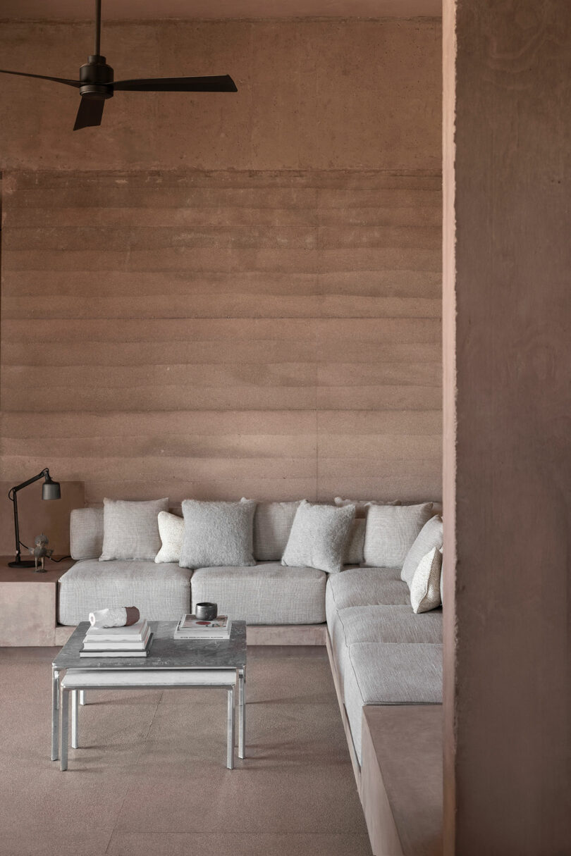 Modern living room with minimalist design, featuring a gray sectional sofa, a small coffee table with books and a cup, a floor lamp, and a ceiling fan, all set against a textured wall.