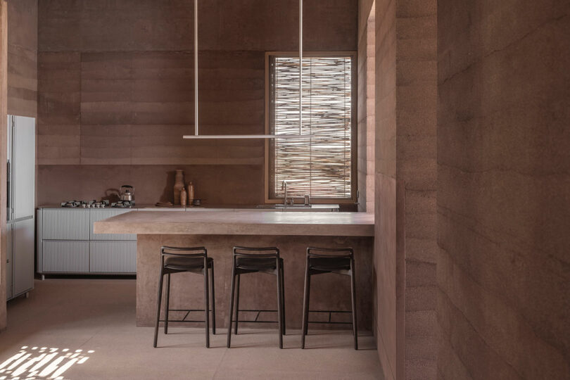 Minimalist kitchen with brown walls and three black barstools at a central island. A window with wooden slats allows light in. The counter on the left holds pottery and kitchen appliances.