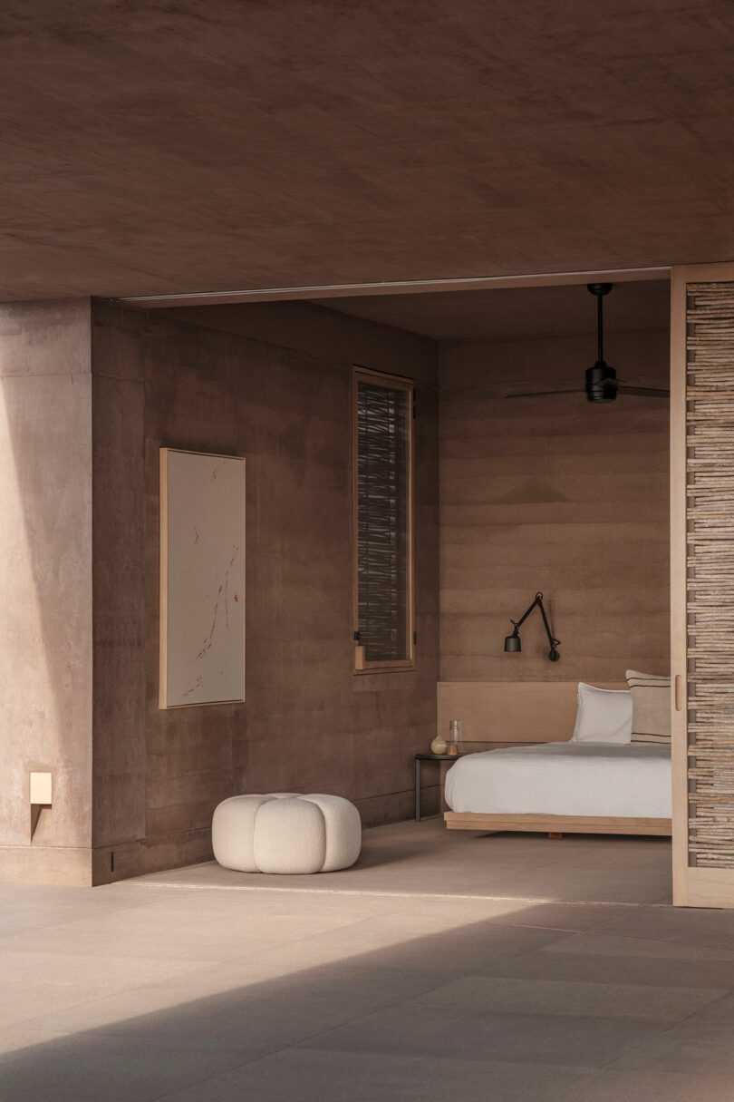 Minimalist bedroom with a wooden bed, white bedding, wall lamp, and a small side table. A round, cushioned stool sits nearby. The walls and ceiling are made of textured, brown material.