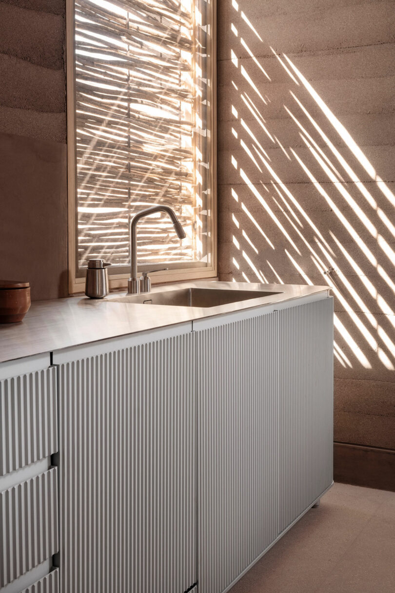 Modern kitchen with a ribbed design cabinet, sleek faucet, and sunlight streaming through a slatted window, casting shadows on the wall.
