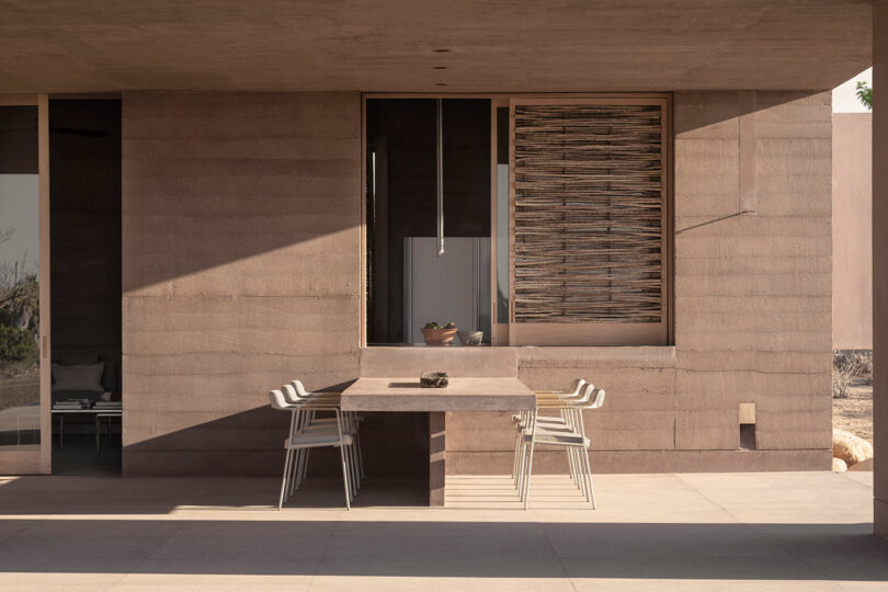 Outdoor patio with a modern concrete table and four chairs, set against a building with stacked wood and a large window.