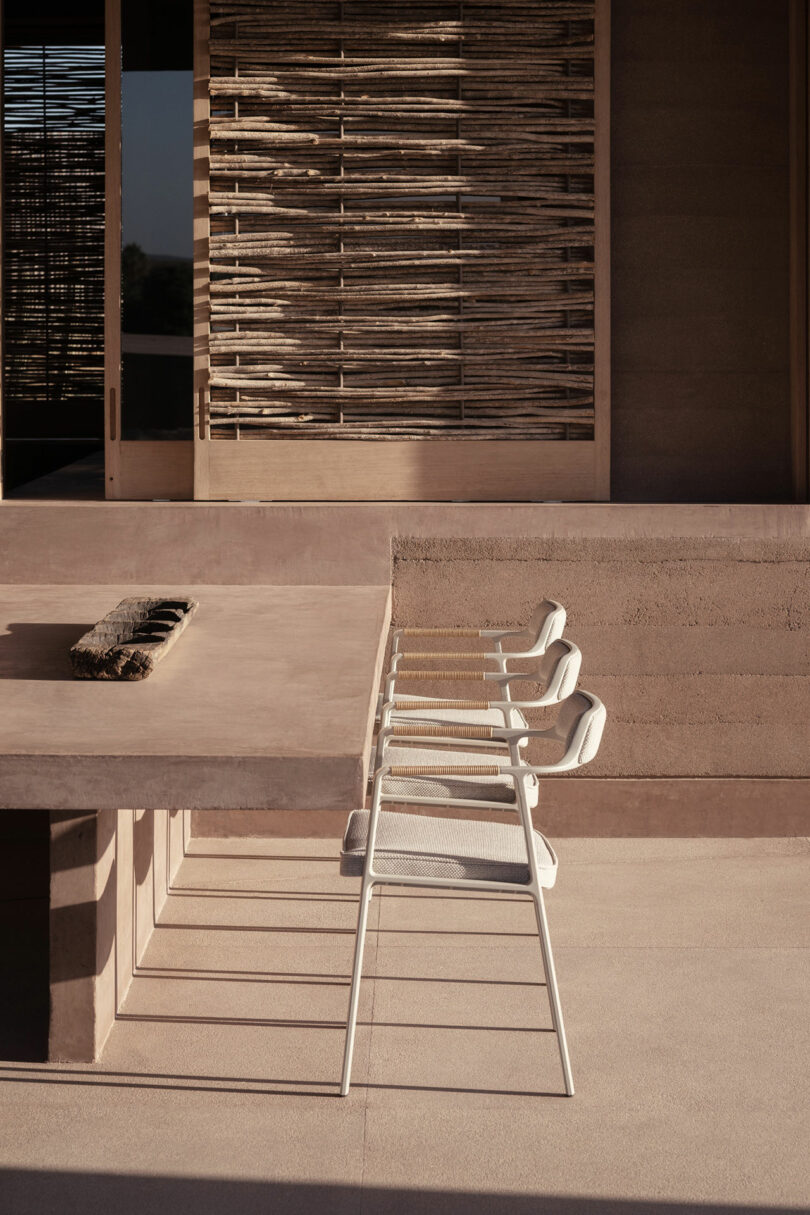 Four white chairs line a beige outdoor table on a sunlit patio with rustic wooden paneling and modern architecture.