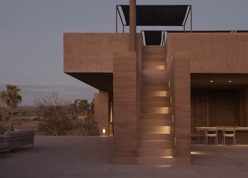Modern concrete building exterior with a central staircase lit from within, surrounded by a patio with outdoor furniture and desert landscape in the background.