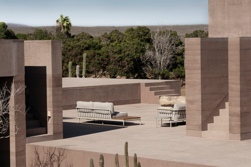 Modern outdoor patio with minimalist beige couches on a spacious terrace surrounded by desert landscape and cacti. Steps lead to differentiated levels.