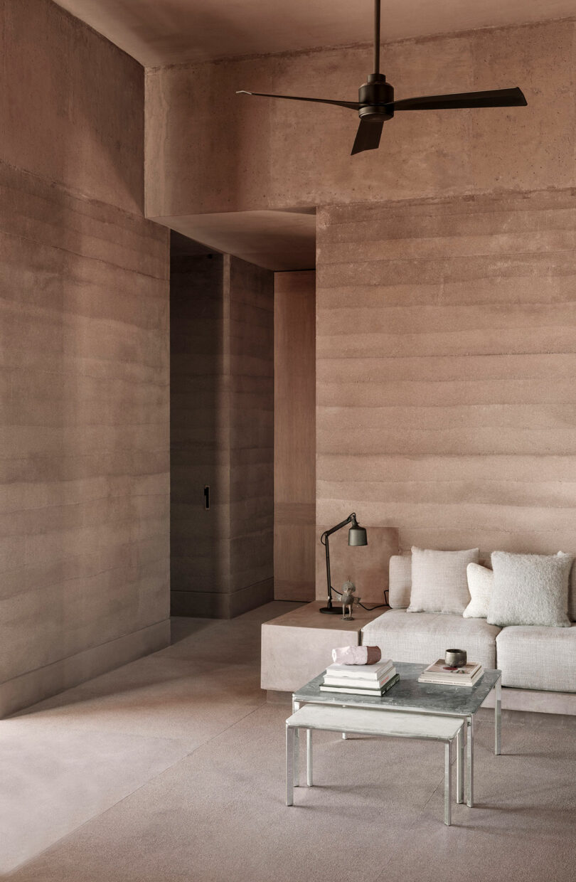 Modern minimalist living room with beige textured walls, a gray sofa, glass coffee table with books, and a black ceiling fan.
