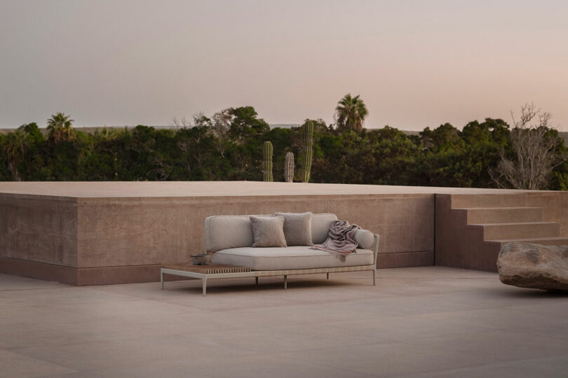 Outdoor patio scene with a modern, minimalist sofa adorned with cushions and a blanket. It faces a lush garden with cacti and trees. The setting is serene, under a clear sky at dusk.