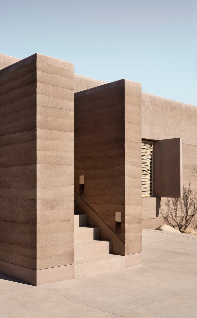 Minimalist desert house exterior with textured brown walls and geometric shapes, including a staircase and a rectangular window.
