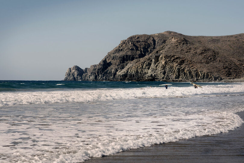 A sandy beach with gentle waves and two people entering the water. A rugged hill rises in the background under a clear blue sky.